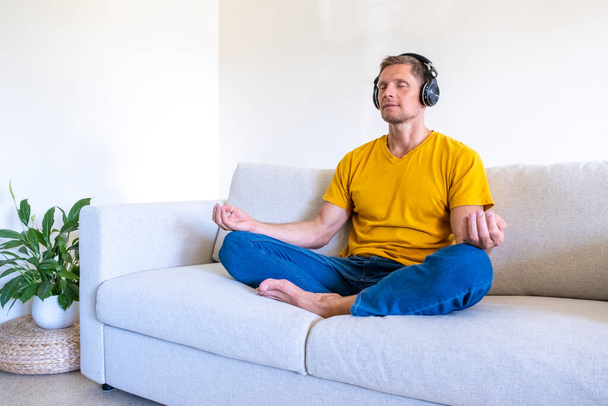 Красивый мужчина с закрытыми глазами сидит в наушниках на диване в позе лотоса и медитирует дома - Фото, изображение