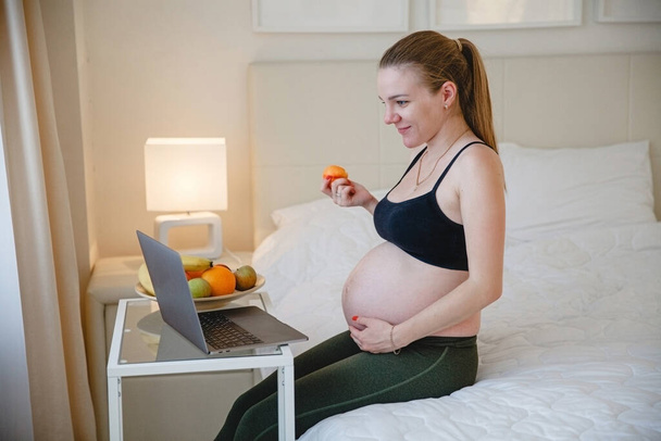 A young blonde pregnant woman is sitting on the bed and eating fruit while looking at her laptop - Zdjęcie, obraz