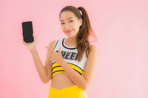 Portrét krásná mladá asijská žena roztleskávačka s chytrým mobilním telefonem na růžové izolované pozadí - Fotografie, Obrázek