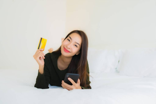 Πορτρέτο όμορφη νεαρή Ασιάτισσα γυναίκα με έξυπνο κινητό τηλέφωνο και πιστωτική κάρτα για online αγορές από το κρεβάτι - Φωτογραφία, εικόνα