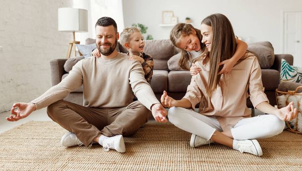 Щаслива сім'я: спокійні молоді батьки сидять на підлозі в позі лотоса і роздумують, поки діти розважаються і обіймають їх вдома
 - Фото, зображення