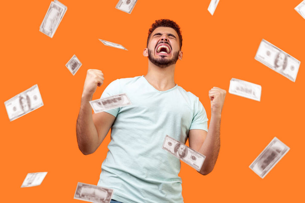 Money rain, winner and rich. Bottom view of ecstatic motivated man standing with raised fists and shouting for joy, winner excited for success. dollars falling. indoor isolated on orange background - Foto, Bild