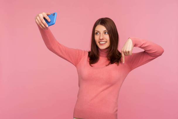 Young woman blogger in pink sweater recording video message to her admirers on smartphone asking to subscribe and rate her posts in social network. Indoor studio shot isolated on pink background - Фото, зображення