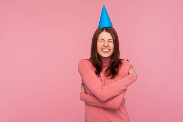 Extremely happy satisfied woman in pink sweater with party cap on head hugging herself congratulating with birthday, excited with party. Indoor studio shot isolated on pink background - Fotó, kép