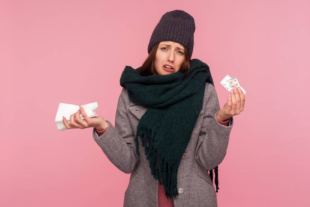 Unhappy frustrated sick woman in warm scarf and hat holding medicine and napkin in hands, stressed because of health problems. Indoor studio shot isolated on pink background - Foto, Bild