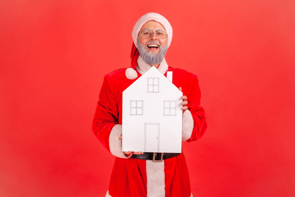Positive smiling man in santa claus costume holding paper house in hands looking at camera with excited expression, dreams comes true on holidays. Indoor studio shot isolated on red background - Fotografie, Obrázek