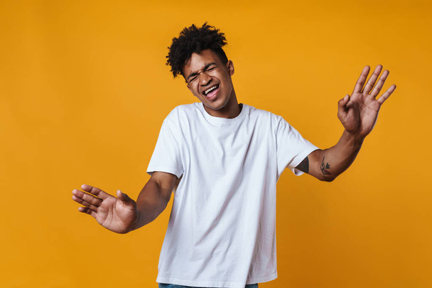 Image of happy african american guy singing and dancing on camera isolated over yellow wall - Φωτογραφία, εικόνα