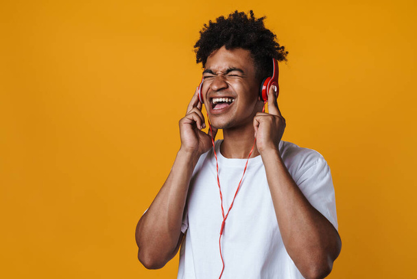 Image of excited african american guy listening music with headphones and singing isolated over yellow wall - Photo, image