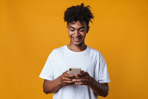 Image of happy african american guy smiling and using mobile phone isolated over yellow wall - Foto, Bild