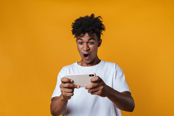 Image of excited african american guy playing online game on cellphone isolated over yellow wall - Foto, imagen