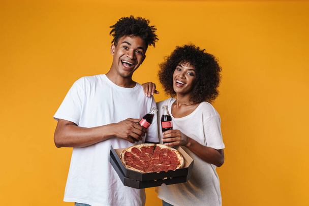 Image of delighted african american couple eating pizza and drinking soda isolated over yellow wall - Фото, зображення