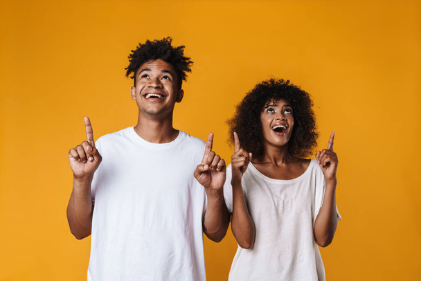 Image of excited african american couple looking and pointing fingers upward isolated over yellow wall - Фото, изображение