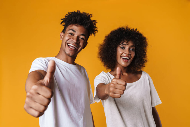 Image of excited african american couple smiling and showing thumbs up isolated over yellow wall - Foto, afbeelding