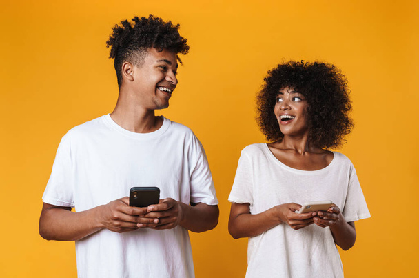 Photo of joyful african american people laughing and using cellphones isolated over yellow background - Foto, imagen