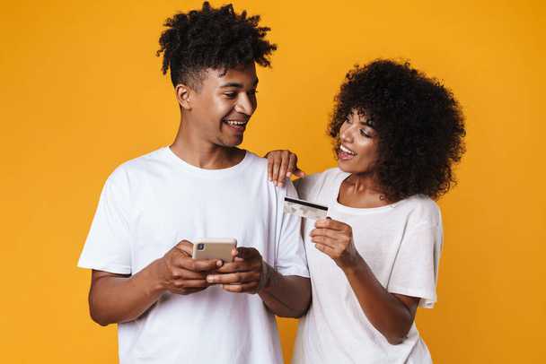 Image of smiling african american couple holding credit card and smartphone isolated over yellow wall - Foto, Imagen