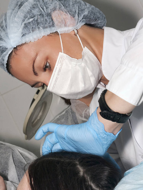 Hautarzt Kosmetologe besucht Patientin vor Schönheitsoperation oder Gesichtsstraffung, schöne Ärztin, Kleid mit OP-Mütze und Maske mit OP-Handschuhen beobachtet die Patientenhaut - Foto, Bild