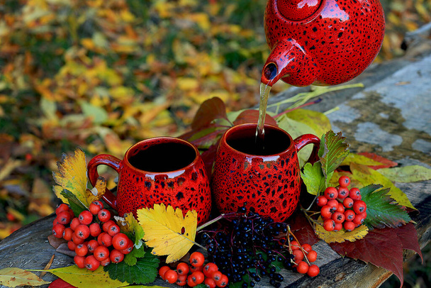 Deux belles tasses rouges et une théière sur un banc en bois dans le parc sur un fond de feuilles d'automne colorées, de baies de rowan rouges et de sureau noir. Le thé chaud est versé de la théière. - Photo, image