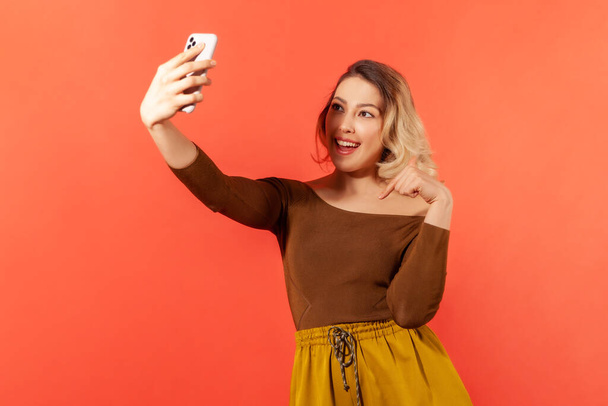 Pretty female blogger recording video on smartphone asking to rate and subscribe on her web page, popularity. Indoor studio shot isolated on orange background - Fotó, kép