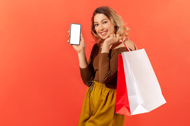 Happy satisfied young woman holding paper bags showing smartphone with white screen, mobile application for shopping online. Indoor studio shot isolated on orange background - Foto, Bild