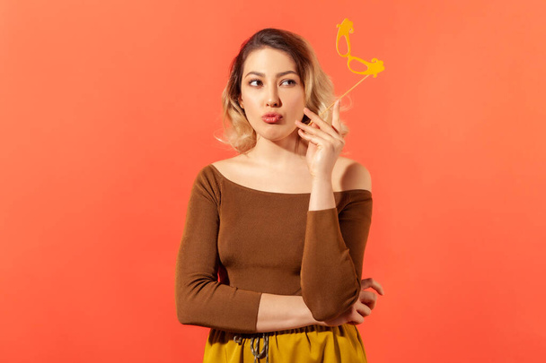 Attractive thoughtful woman holding paper mask on stick contemplating about her costume at fancy party, dreaming. Indoor studio shot isolated on orange background - Photo, image