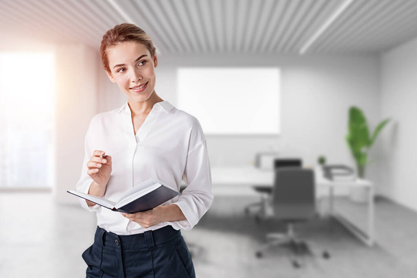 Portrait of smiling young businesswoman standing with notebook and taking notes in blurry office. Toned image - Photo, image