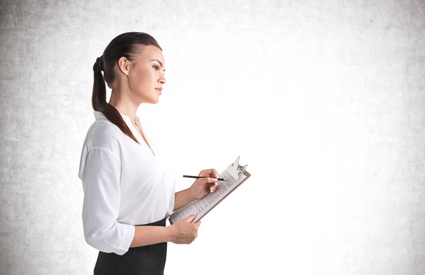 Side portrait of serious young European businesswoman standing with clipboard over concrete background. Mock up - Photo, Image