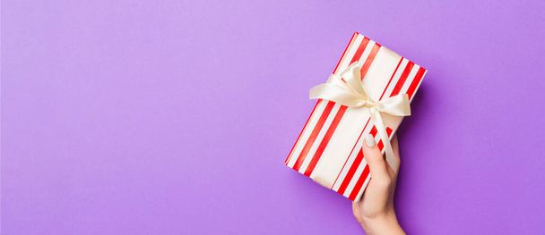 Flat lay of woman hands holding gift wrapped and decorated with bow on purple background with copy space. Christmas and holiday concept. - Фото, зображення