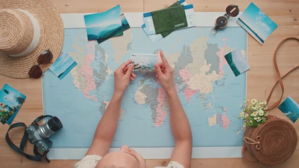 Horizontal top-view flat lay footage of unrecognizable woman holding printed photos of beautiful landmarks in hands laying them on world map - Filmagem, Vídeo