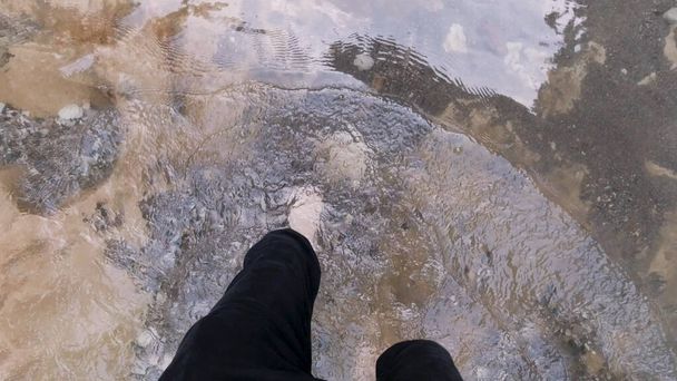 Pies de hombre con piernas peludas cruzando el frío arroyo de montaña. Clip. Vista superior de las piernas desnudas masculinas caminando a través del río frío con fondo pedregoso, concepto de senderismo. - Foto, imagen
