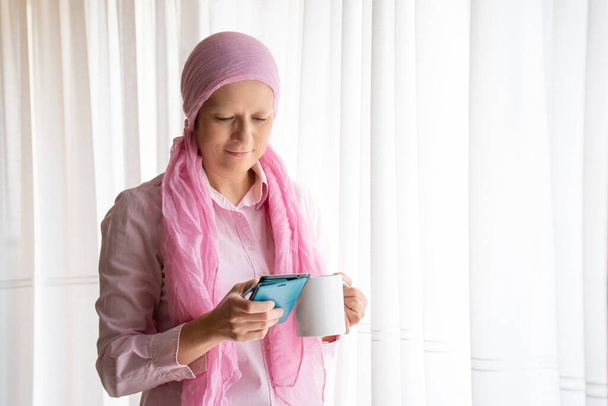 Жінка з раком і рожевим хусткою дивиться на мобільний телефон і посміхається біля вікна
 - Фото, зображення
