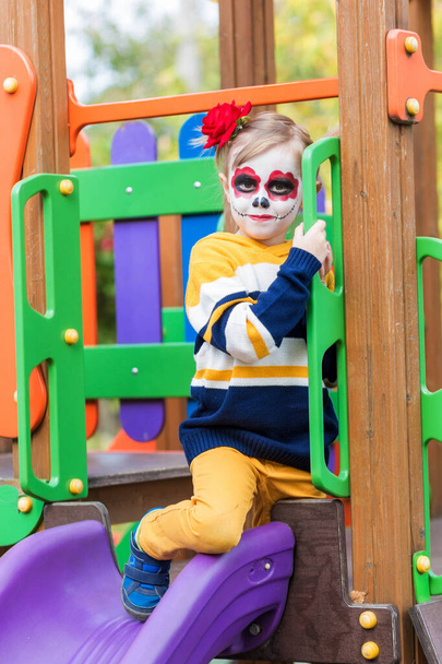 A little preschool girl with Painted Face, rides a slide on the playground, celebrates Halloween or Mexican Day of the Dead. - Фото, зображення