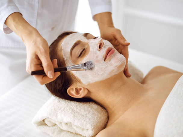 Beautiful brunette woman enjoying applying cosmetic mask with closed eyes. Relaxing treatment in medicine and spa center concepts - Foto, afbeelding
