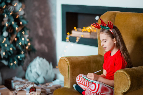 A cute girl in a Santa hat writes a letter to Santa near the Christmas tree. Happy childhood, a time for fulfilling desires. Merry Christmas. - Photo, Image