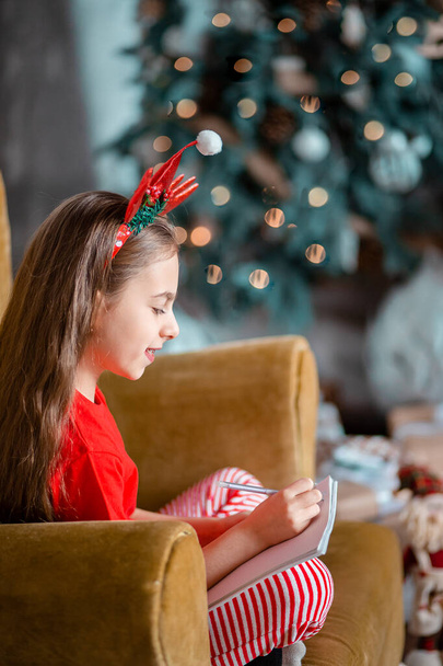A cute girl in a Santa hat writes a letter to Santa near the Christmas tree. Happy childhood, a time for fulfilling desires. Merry Christmas. - Foto, Bild
