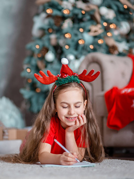 A cute girl in a Santa hat writes a letter to Santa near the Christmas tree. Happy childhood, a time for fulfilling desires. Merry Christmas. - Fotografie, Obrázek