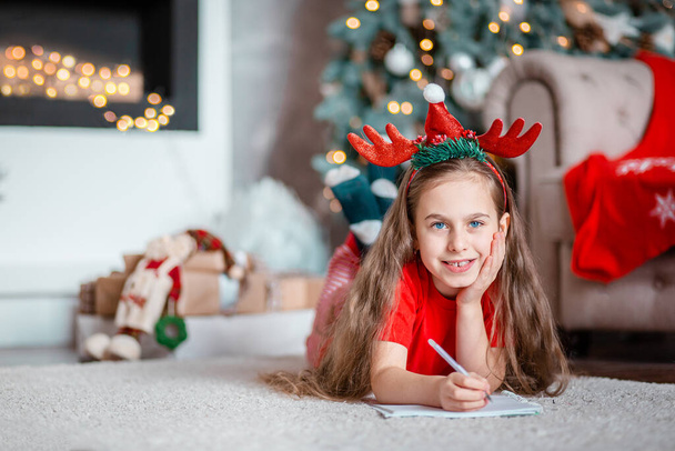 A cute girl in a Santa hat writes a letter to Santa near the Christmas tree. Happy childhood, a time for fulfilling desires. Merry Christmas. - Foto, afbeelding