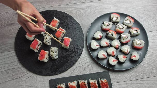 La mano femenina toma comida con palillos. Composición de comida japonesa. Varios tipos de sushi colocados en tablero de piedra negra. Ensalada de kimchi picante, sopa de wonton, palillos y salsa de soja. - Foto, Imagen