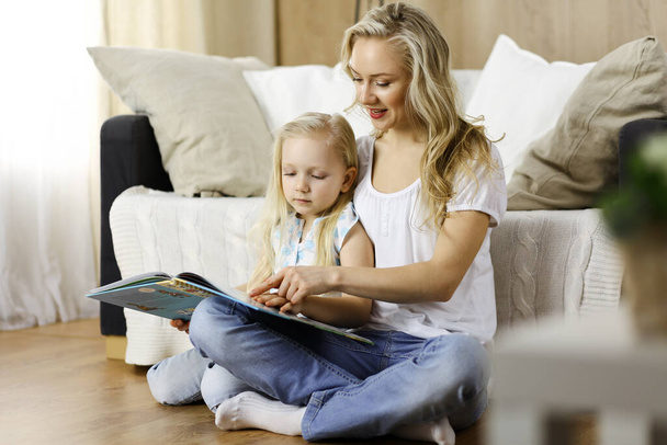 Happy family. Blonde young mother reading a book to her cute daughter while sitting at wooden floor. Motherhood concept - Foto, Imagem