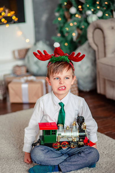 Angry upset little boy with a gift, toy train, under the Christmas tree on a New Year's morning. A time of miracles and fulfillment of desires. Merry Christmas. - Фото, зображення