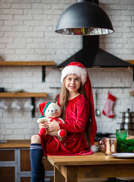 Frohe Weihnachten. Nettes kleines Mädchen mit Weihnachtsmütze hält einen Teddybär in der Küche und wartet auf den Urlaub. Eine Zeit der Wunder und der Erfüllung von Wünschen. - Foto, Bild