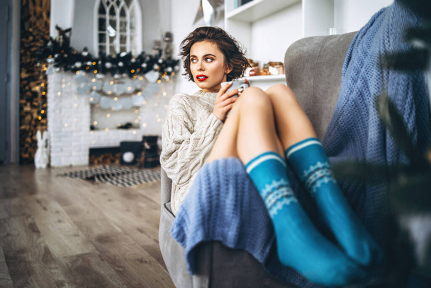 Pretty and happy brunette sitting in warm sweater on the chair, holding cup of hot beverage with christmas lights behind - Foto, Bild