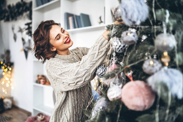 Christmas, New Year. Pretty and happy brunette in warm sweater decorating christmas tree at home - Foto, Imagem