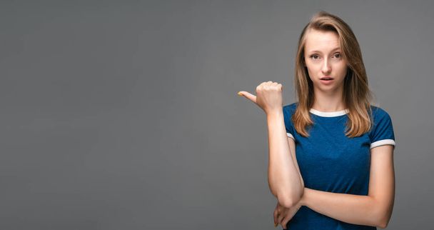Young woman with blonde straight hair over isolated gray background pointing finger to the side. Presenting your product. Expressive facial. Copy space for your text - Fotografie, Obrázek