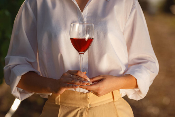Mujer viticultora degustación de vino tinto de una copa en un viñedo. viñedos fondo al atardecer. Macro disparo de una mano sommelier que sostiene la copa de vino - Foto, imagen