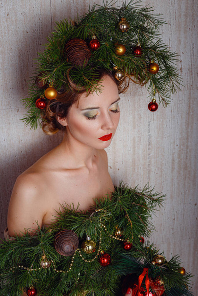 Beautiful happy young lady with a Christmas pine wreath on her head and fashione xmas tree dress, decorated with Christmas balls, berries and snowflakes. New Year Concept - Фото, зображення