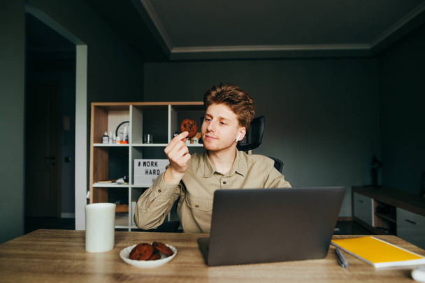 Funny young man in a shirt and wireless headphones works at home on a laptop in the workplace and eats cookies with tea for lunch. Freelancer eats cookies. - Photo, Image