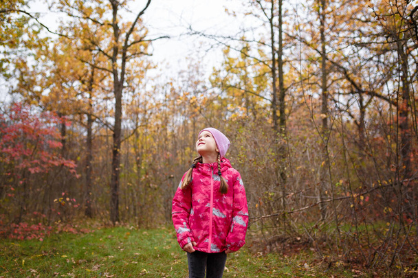 Маленькая девочка в розовой куртке и вязаной шапке стоит в осеннем лесу - Фото, изображение