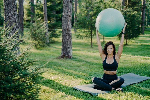 Brunette slim woman lifts fitness ball over head, sits in lotus pose on karemat, dressed in active wear, practices yoga outside, breathes fresh air in forest. Fit lady exercises with gymnastic ball - Photo, Image