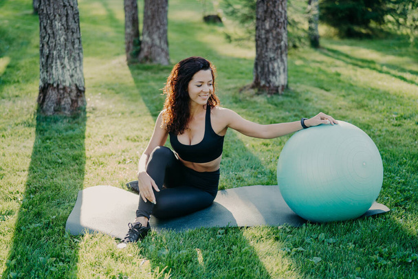 Outdoor shot of active sporty woman takes break after physical exercises, poses on fitness mat with fitball, enjoys fresh air in forest, has perfect body shape uses sport equipment. Gymnastics concept - Foto, immagini