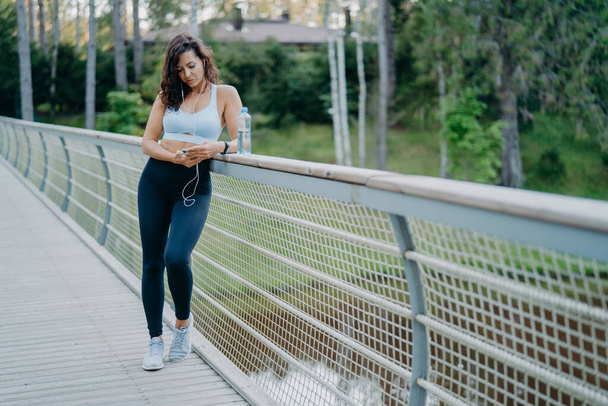 Sporty slim European woman in active wear checks notification on mobile phone, listens music from playlist uses earphones, poses on bridge against nature background. Sport and lifestyle concept - Foto, immagini